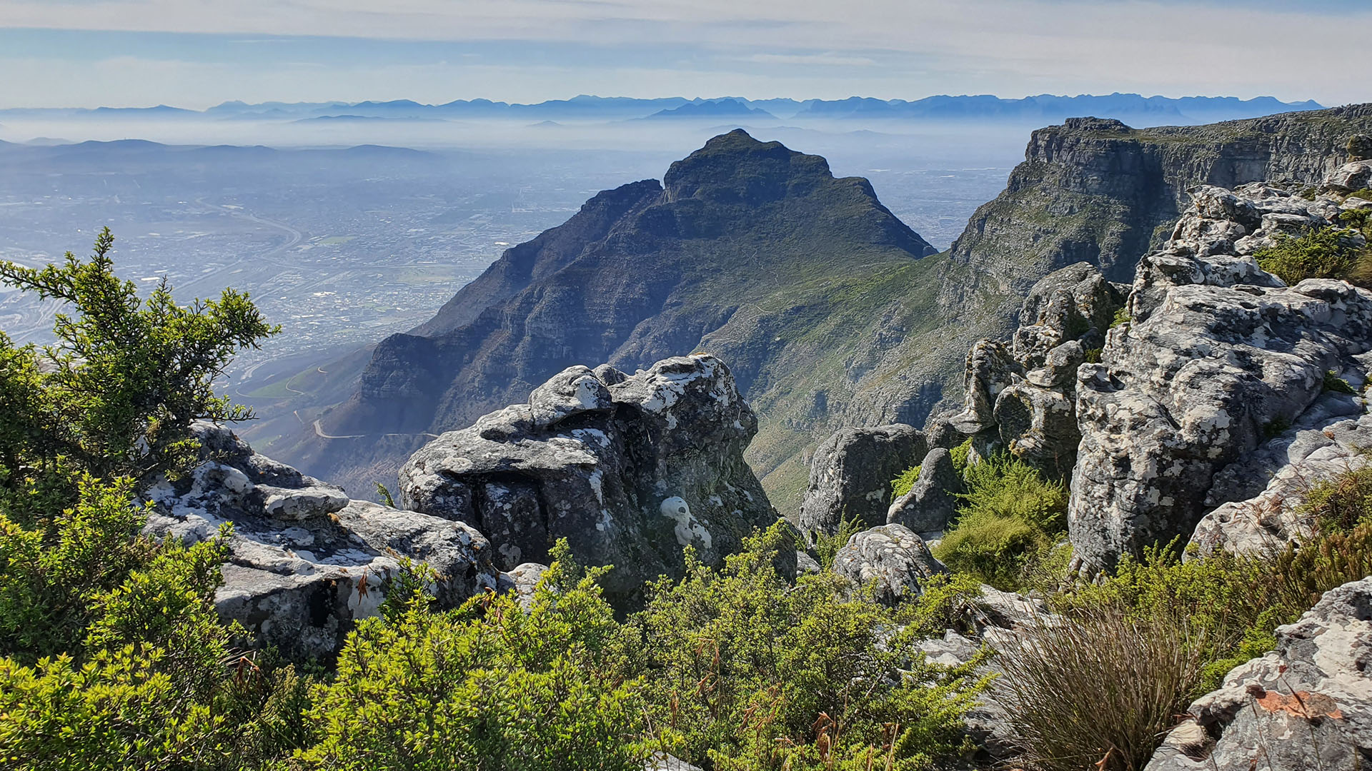 Expansive view from a rocky hiking trail on Table Mountain, looking towards Devil's Peak and the hazy Cape Town suburbs.